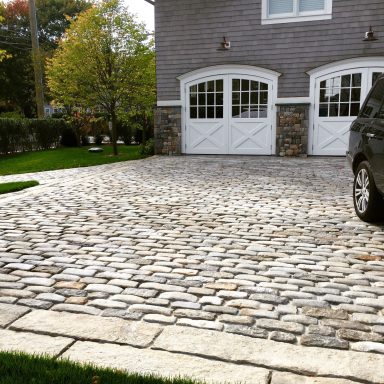 Cobblestone driveway with a black car parked beside two garage doors.